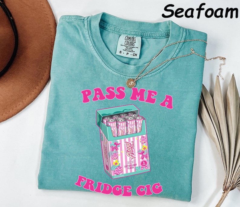 May include: Seafoam green t-shirt with the text "PASS ME A FRIDGE CIG" in pink. The graphic is a box of pink and white cans. A gold necklace is draped on the shirt.