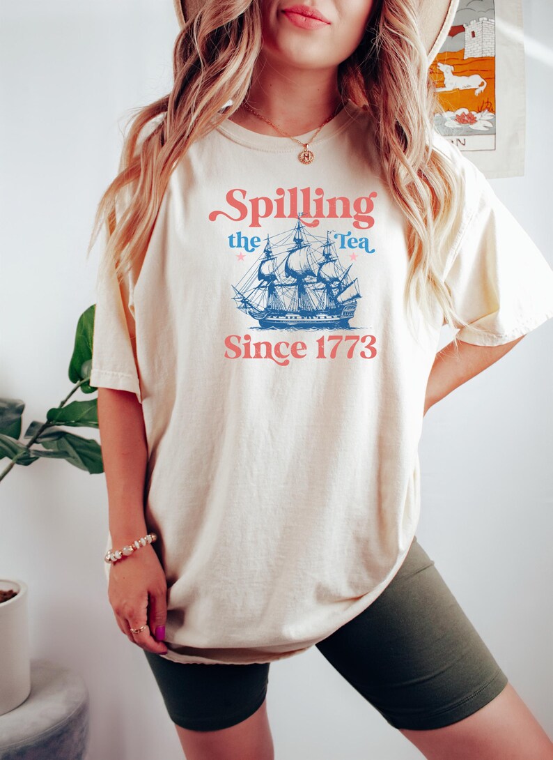May include: Cream-colored t-shirt with the text "Spilling the Tea Since 1773" in pink and blue. The design features a blue illustration of a tall ship. The shirt is a casual, comfortable style.