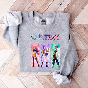 May include: A heather gray sweatshirt featuring the word "HUNTXRX" in a colorful, stylized font. Below the text are three illustrated figures in vibrant outfits. The sweatshirt is folded on a light wood surface.