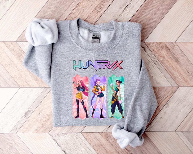 May include: A heather gray sweatshirt featuring the word "HUNTXRX" in a colorful, stylized font. Below the text are three illustrated figures in vibrant outfits. The sweatshirt is folded on a light wood surface.