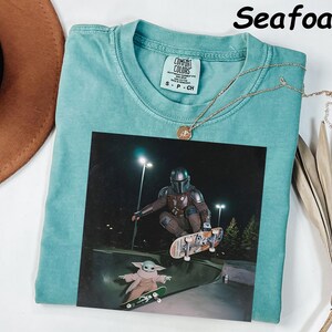 May include: Seafoam green t-shirt featuring a graphic of a character from a science fiction series skateboarding. The graphic includes a character in armour and a small green alien. A gold necklace is draped across the shirt.