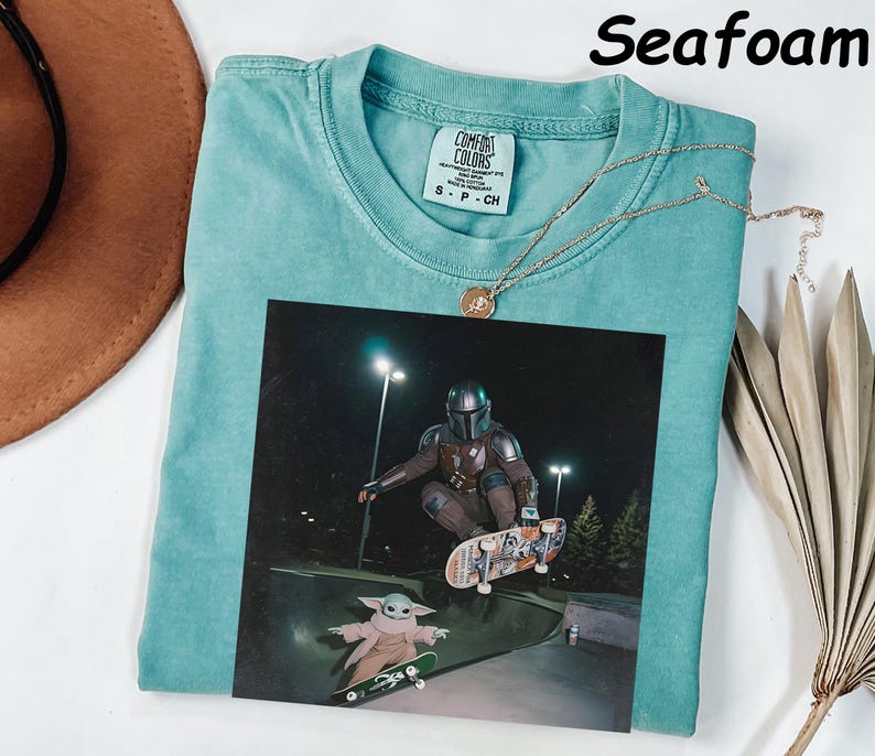 May include: Seafoam green t-shirt featuring a graphic of a character from a science fiction series skateboarding. The graphic includes a character in armour and a small green alien. A gold necklace is draped across the shirt.