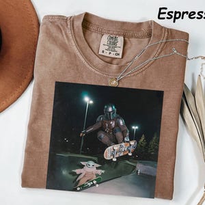 May include: Espresso-coloured t-shirt with a graphic of a character from a popular sci-fi series skateboarding with a small green alien. The shirt has a crew neck and a small tag. A silver necklace with a pendant is draped across the shirt.