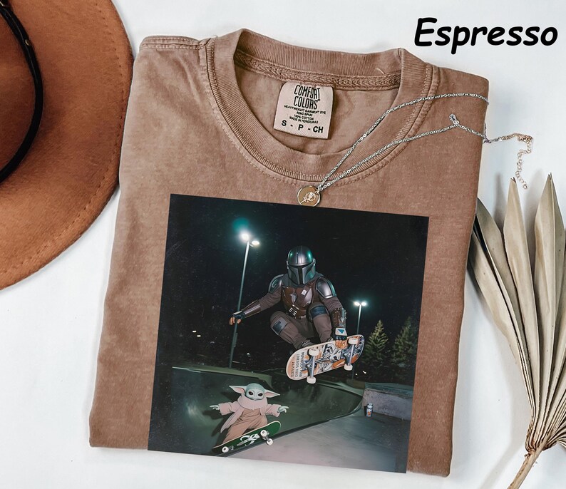 May include: Espresso-coloured t-shirt with a graphic of a character from a popular sci-fi series skateboarding with a small green alien. The shirt has a crew neck and a small tag. A silver necklace with a pendant is draped across the shirt.