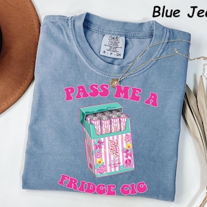 May include: Blue jean colored t-shirt with the text "PASS ME A FRIDGE CIG" in pink. The shirt features a graphic of a cigarette box with pink cans inside. A gold necklace is draped across the shirt.