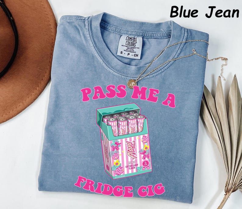 May include: Blue jean colored t-shirt with the text "PASS ME A FRIDGE CIG" in pink. The shirt features a graphic of a cigarette box with pink cans inside. A gold necklace is draped across the shirt.