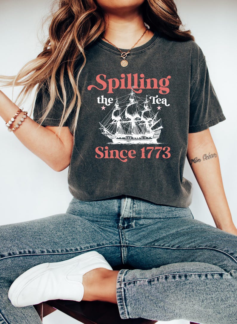 May include: Dark gray t-shirt with the text "Spilling the Tea Since 1773" in a vintage style. The design includes a ship illustration. The shirt is paired with blue jeans and white sneakers.