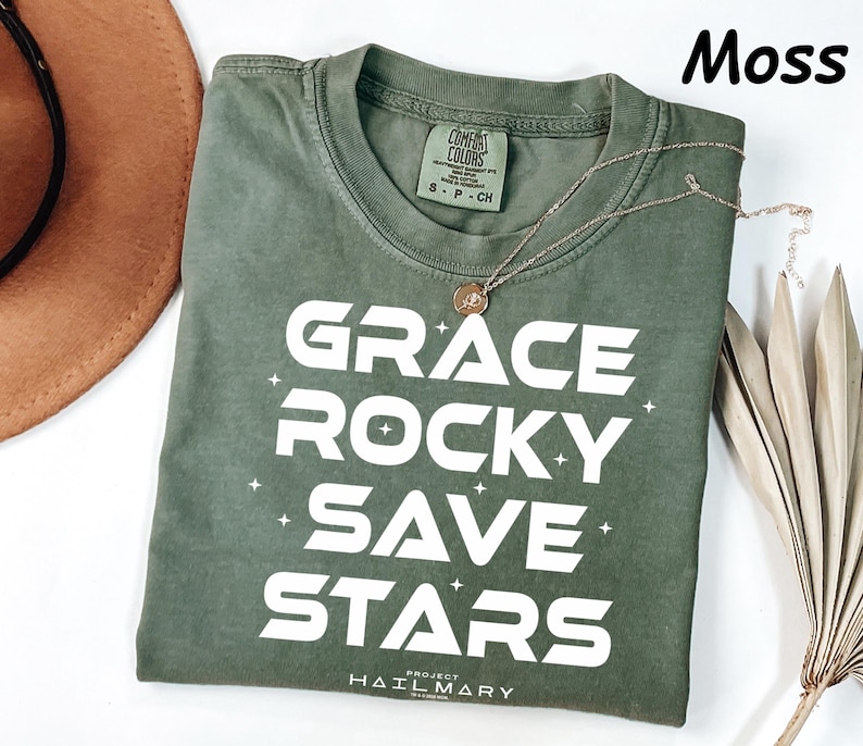 May include: A moss green t-shirt with the words "GRACE ROCKY SAVE STARS" in white block letters. The shirt has a crew neck and short sleeves. A gold necklace with a pendant is draped across the shirt. The text "PROJECT HAIL MARY" is printed at the bottom.