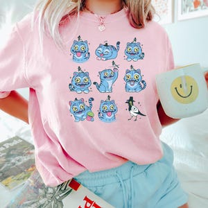 May include: A light pink sweatshirt featuring a repeating pattern of blue cartoon cats with various expressions and accessories. The cats have yellow eyes and are set against a light pink background. The person is holding a white mug with a yellow smiley face.