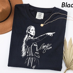 May include: A black short-sleeved t-shirt featuring a white graphic print of a basketball player pointing, with the number 8 and a signature. The shirt has a Comfort Colors brand label at the neck and the text Black is printed in the top right corner.