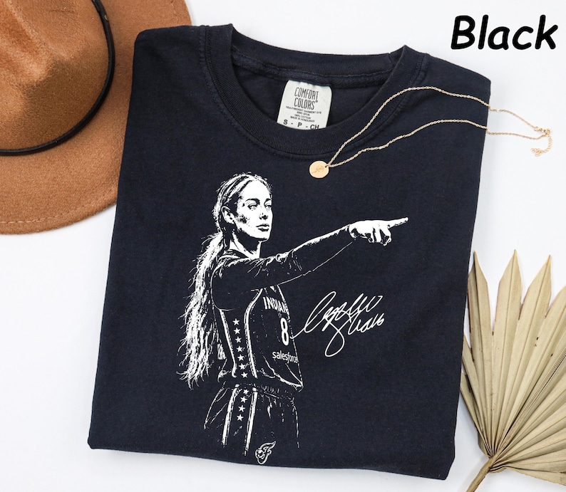 May include: A black short-sleeved t-shirt featuring a white graphic print of a basketball player pointing, with the number 8 and a signature. The shirt has a Comfort Colors brand label at the neck and the text Black is printed in the top right corner.