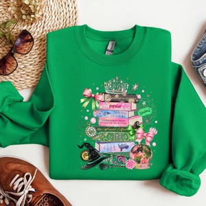May include: A green sweatshirt featuring a whimsical design of stacked books, a witch's hat, a crystal ball, and a crown. The books have titles like "Popular" and "Defying Gravity." Pink bows and pearls add decorative accents to the design.