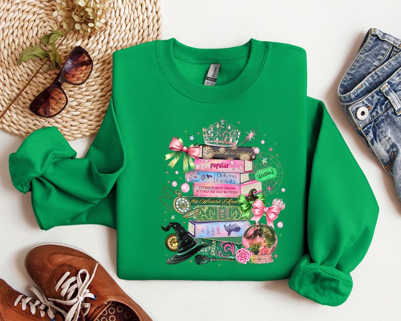 May include: A green sweatshirt featuring a whimsical design of stacked books, a witch's hat, a crystal ball, and a crown. The books have titles like "Popular" and "Defying Gravity." Pink bows and pearls add decorative accents to the design.