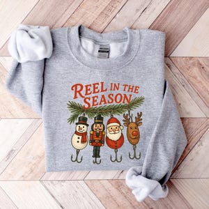 May include: A heather gray sweatshirt with the text "REEL IN THE SEASON" in red. Below the text are four Christmas-themed fishing lures: a snowman, a nutcracker, Santa Claus, and a reindeer. The sweatshirt is folded on a wooden surface.