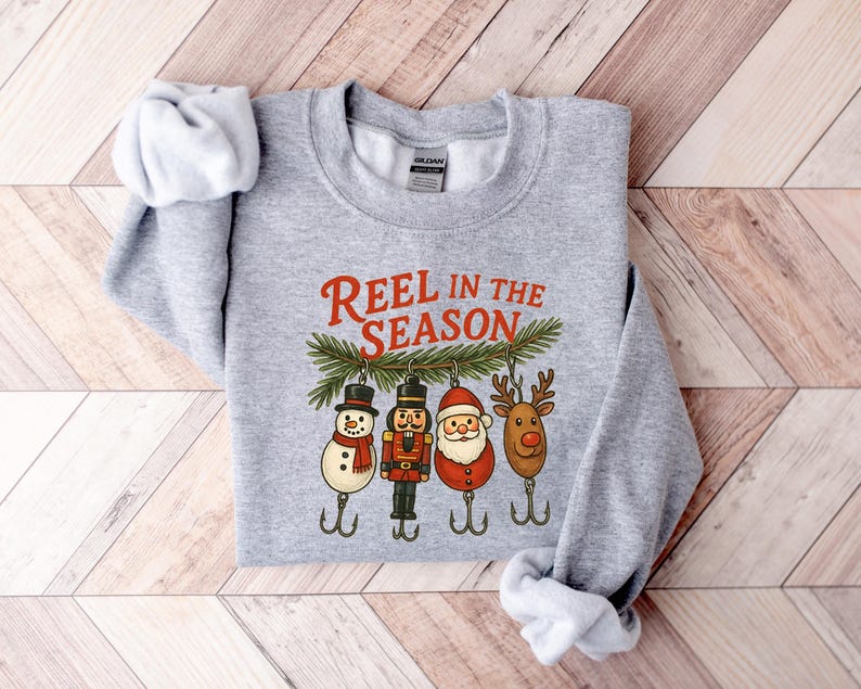 May include: A heather gray sweatshirt with the text "REEL IN THE SEASON" in red. Below the text are four Christmas-themed fishing lures: a snowman, a nutcracker, Santa Claus, and a reindeer. The sweatshirt is folded on a wooden surface.