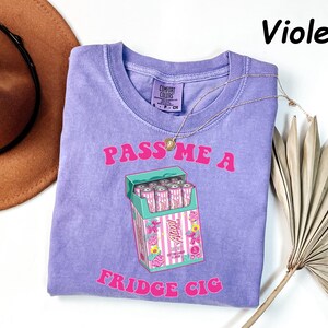 May include: Violet-colored t-shirt with the text "PASS ME A FRIDGE CIG" in pink. The graphic features a cartoon cigarette box filled with pink cans. A gold necklace and a brown hat are also visible.