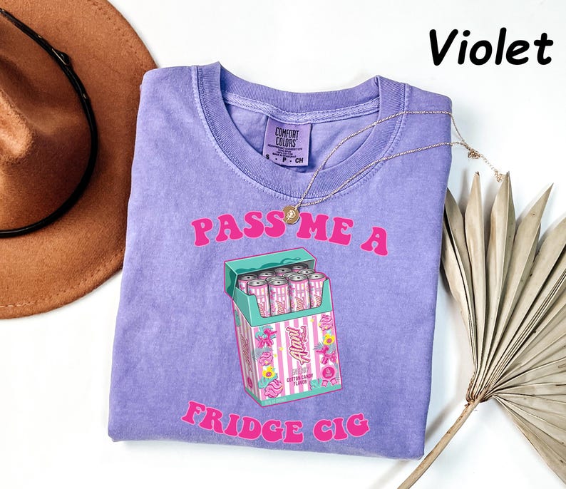 May include: Violet-colored t-shirt with the text "PASS ME A FRIDGE CIG" in pink. The graphic features a cartoon cigarette box filled with pink cans. A gold necklace and a brown hat are also visible.