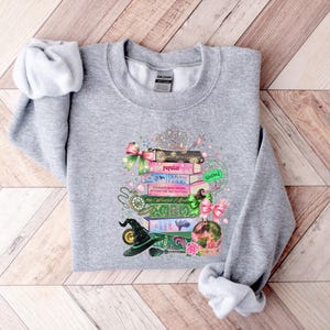 May include: A heather gray sweatshirt featuring a whimsical design of stacked books, a witch's hat, and a crystal ball. The books have titles like "Popular" and "The Wizard's Guide". Pink bows and a silver tiara add decorative accents to this cozy top.