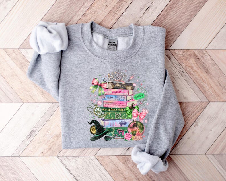 May include: A heather gray sweatshirt featuring a whimsical design of stacked books, a witch's hat, and a crystal ball. The books have titles like "Popular" and "The Wizard's Guide". Pink bows and a silver tiara add decorative accents to this cozy top.