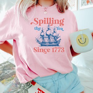 May include: A light pink t-shirt with the text "Spilling the Tea Since 1773" and a blue illustration of a tall ship. The person is holding a white mug with a yellow smiley face.