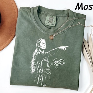 May include: A moss green short-sleeve t-shirt featuring a white graphic print of a basketball player pointing, with the number 8 and a signature. The shirt has a crew neckline and a Comfort Colors brand label on the inside collar with size S-CH.