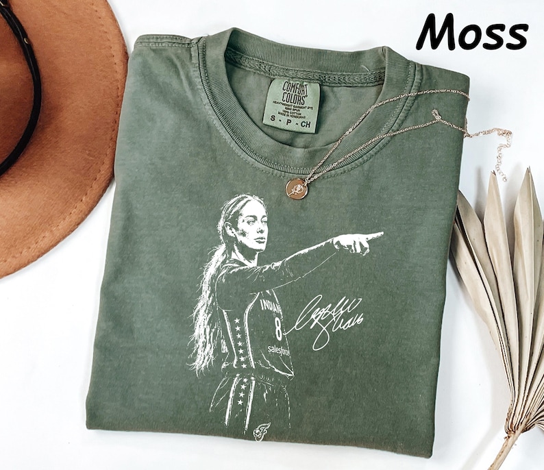 May include: A moss green short-sleeve t-shirt featuring a white graphic print of a basketball player pointing, with the number 8 and a signature. The shirt has a crew neckline and a Comfort Colors brand label on the inside collar with size S-CH.