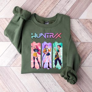 May include: Olive green sweatshirt with the word "HUNTRAX" in a colorful font. Below the text are three anime-style illustrations of women in action poses, each in a different outfit. The sweatshirt has a crew neck and long sleeves.