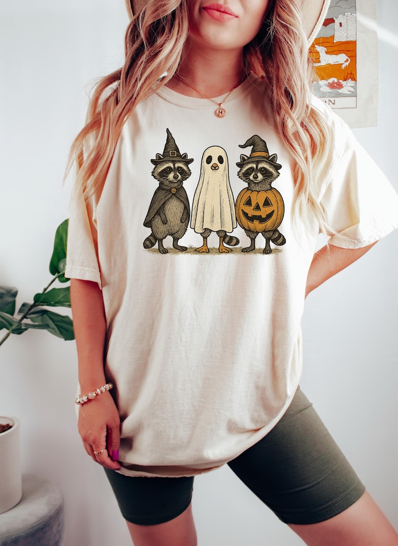 May include: Cream-colored t-shirt featuring a Halloween-themed graphic. The graphic depicts three cartoon characters: a raccoon in a witch hat, a ghost, and a raccoon inside a carved pumpkin. The design is in shades of brown, black, orange, and white.