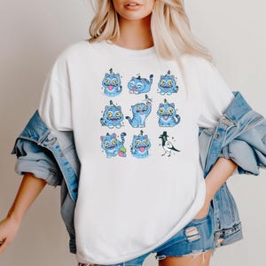 May include: White t-shirt featuring a repeating pattern of blue cartoon cats with various expressions and poses. A black and white bird is also included in the design. The cats have yellow eyes and are wearing small hats. The shirt is worn by a person.