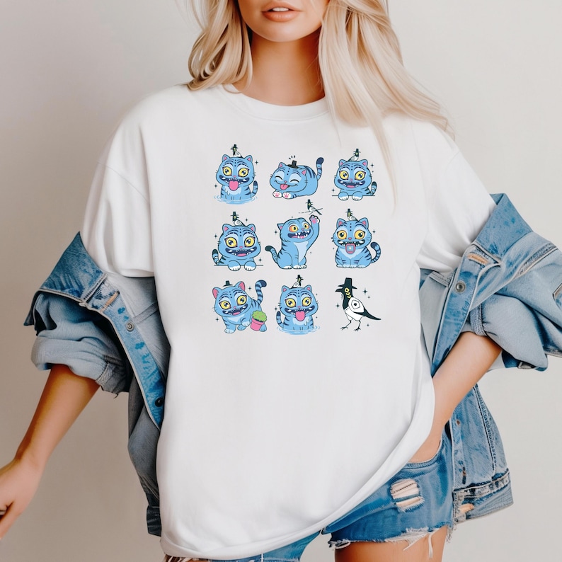 May include: White t-shirt featuring a repeating pattern of blue cartoon cats with various expressions and poses. A black and white bird is also included in the design. The cats have yellow eyes and are wearing small hats. The shirt is worn by a person.