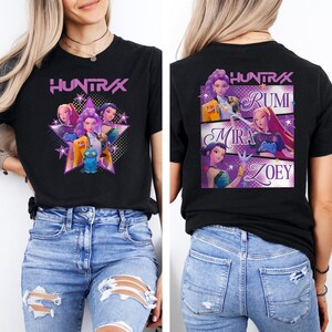 May include: Black t-shirt featuring the text "HUNTRAX" in purple, with a graphic of several animated characters. The back of the shirt displays the names Rumi, Mira, and Zoey, also with character graphics. A stylish tee.