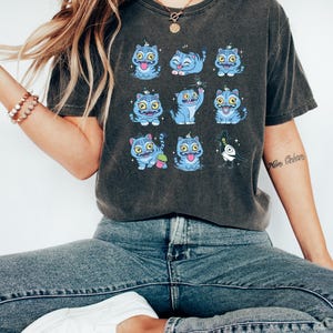 May include: Dark gray t-shirt featuring a grid of blue cartoon cats with various expressions. The cats have yellow eyes and are decorated with small blue hats. The shirt is worn with blue jeans and white sneakers.