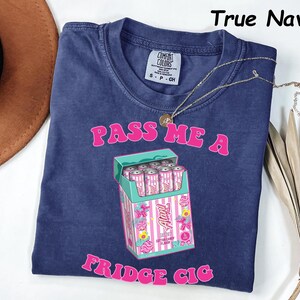 May include: A navy blue t-shirt with the text "PASS ME A FRIDGE CIG" in pink. The graphic features a box of pink and white cans. A gold necklace is draped across the shirt.