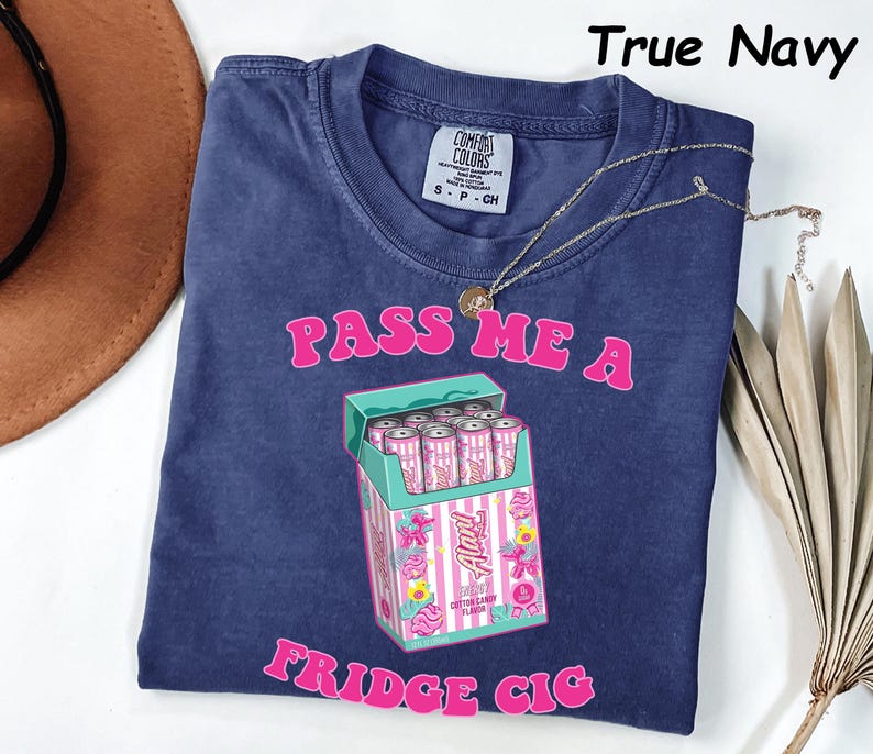 May include: A navy blue t-shirt with the text "PASS ME A FRIDGE CIG" in pink. The graphic features a box of pink and white cans. A gold necklace is draped across the shirt.
