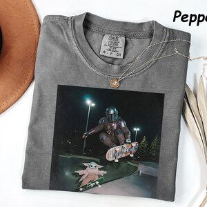 May include: Grey t-shirt with a graphic of a character skateboarding. The image features a character in a helmet and armour, and a smaller character on a skateboard. A silver necklace with a pendant is on the shirt.