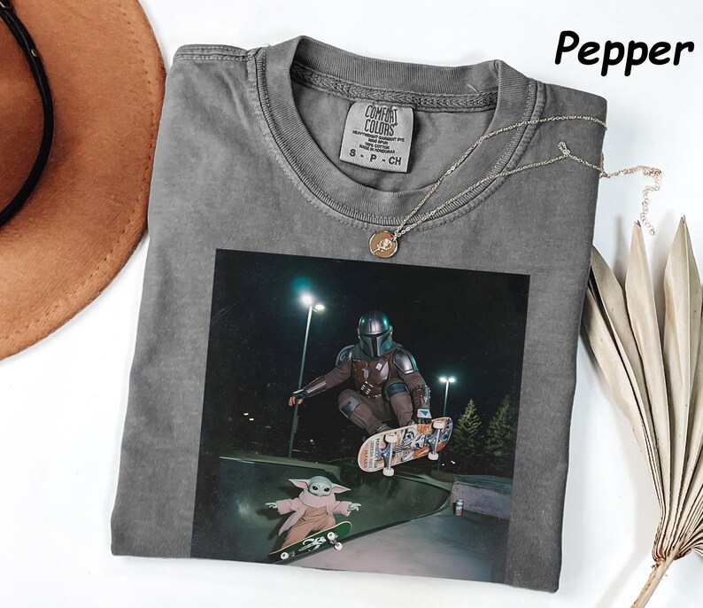 May include: Grey t-shirt with a graphic of a character skateboarding. The image features a character in a helmet and armour, and a smaller character on a skateboard. A silver necklace with a pendant is on the shirt.