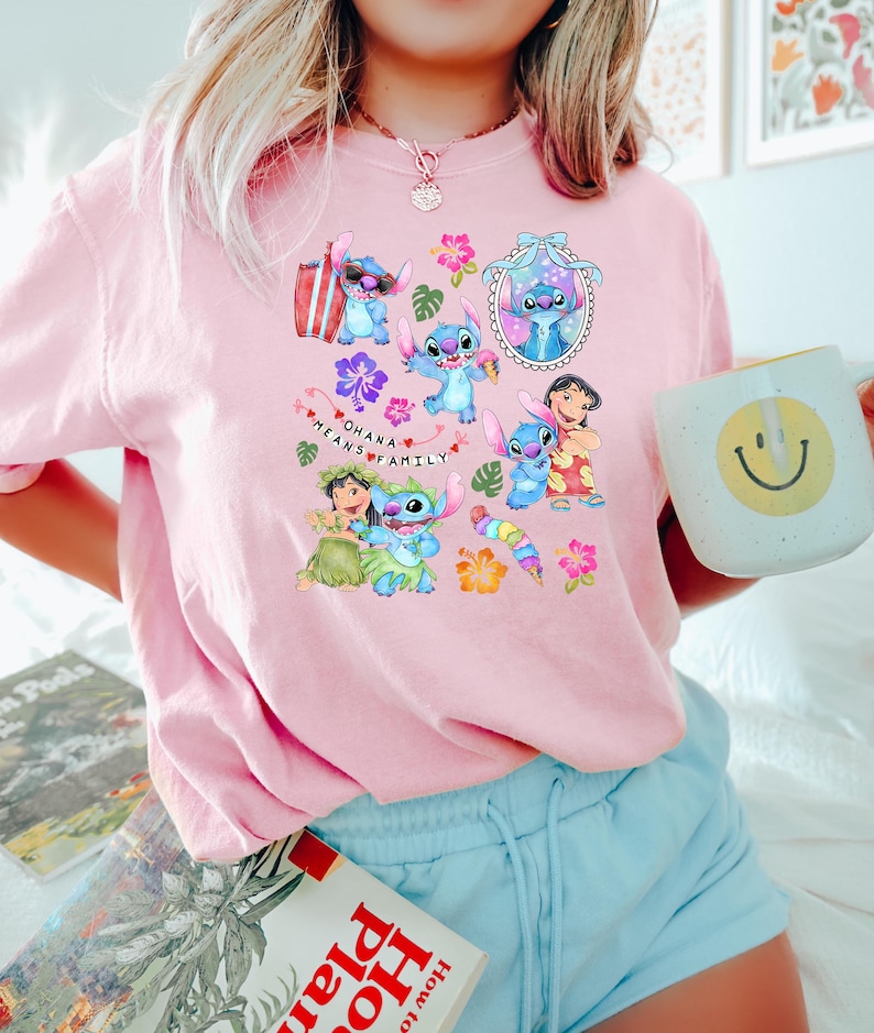 Peut inclure: Un t-shirt rose clair avec un motif color&eacute; de Stitch de Lilo & Stitch, entour&eacute; de fleurs et du texte "Ohana Means Family". Le motif comprend diverses images de Stitch. Une tasse blanche avec un smiley jaune est tenue dans la main.