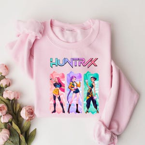 May include: A light pink sweatshirt featuring the word "HUNTRAX" in a colorful font. Below the text are three anime-style characters in colorful outfits. The sweatshirt is made of a soft material and is suitable for casual wear.