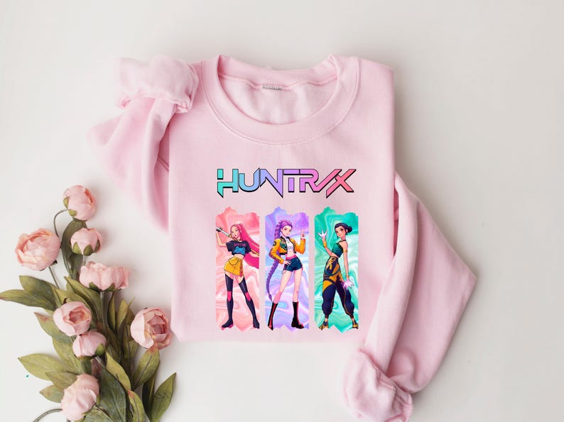 May include: A light pink sweatshirt featuring the word "HUNTRAX" in a colorful font. Below the text are three anime-style characters in colorful outfits. The sweatshirt is made of a soft material and is suitable for casual wear.