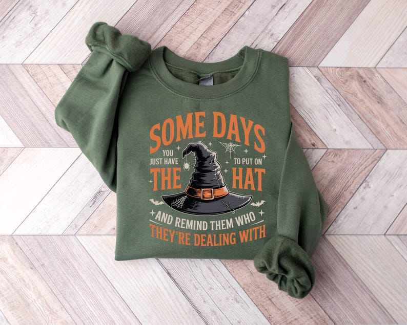 Puede incluir: Una sudadera verde oliva con el texto "SOME DAYS you just have the HAT and remind them who they're dealing with." El gr&aacute;fico presenta un sombrero de bruja negro con una hebilla naranja. La sudadera est&aacute; doblada sobre una superficie de madera.