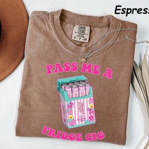 May include: Espresso-colored t-shirt with the text "PASS ME A FRIDGE CIG" in pink. The shirt features a graphic of a cigarette box filled with pink cans. A silver necklace is draped across the shirt.