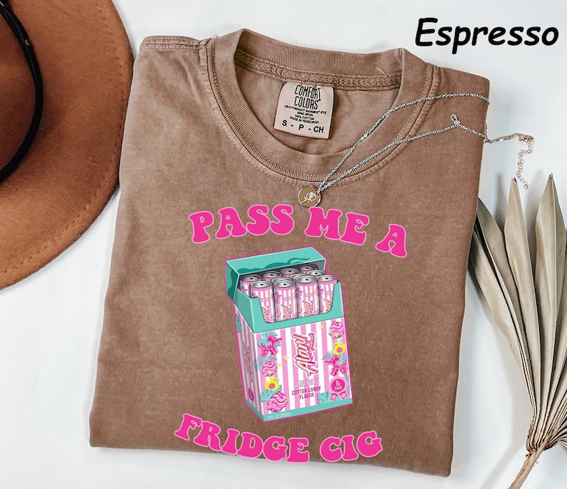 May include: Espresso-colored t-shirt with the text "PASS ME A FRIDGE CIG" in pink. The shirt features a graphic of a cigarette box filled with pink cans. A silver necklace is draped across the shirt.