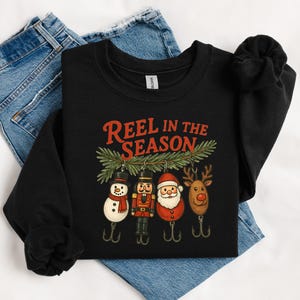 May include: Black sweatshirt with the text "Reel in the Season" in red. The graphic features Christmas-themed fishing lures: a snowman, nutcracker, Santa, and reindeer. The sweatshirt is displayed on a pair of blue jeans.