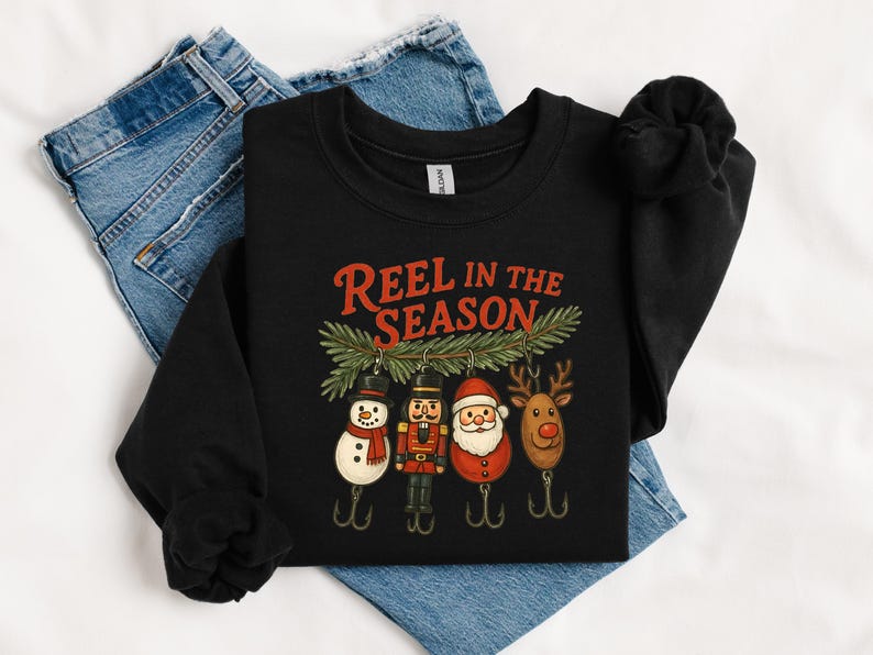 May include: Black sweatshirt with the text "Reel in the Season" in red. The graphic features Christmas-themed fishing lures: a snowman, nutcracker, Santa, and reindeer. The sweatshirt is displayed on a pair of blue jeans.