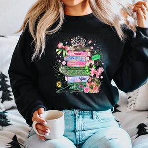 May include: Black sweatshirt with a colorful graphic of stacked books, a crown, and a snow globe. The books have titles like "Popular" and "Defying Gravity." Pink and green bows and a witch's hat add to the whimsical design.