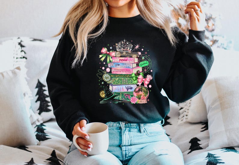 May include: Black sweatshirt with a colorful graphic of stacked books, a crown, and a snow globe. The books have titles like "Popular" and "Defying Gravity." Pink and green bows and a witch's hat add to the whimsical design.