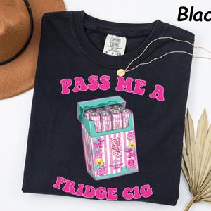 May include: Black t-shirt with the text "PASS ME A FRIDGE CIG" in pink. The shirt features a graphic of a cigarette box with pink and white cans inside. A gold necklace is draped across the shirt. A brown hat is in the upper left corner.