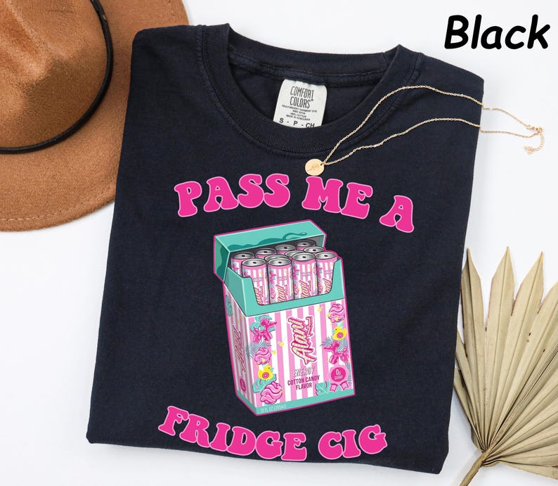 May include: Black t-shirt with the text "PASS ME A FRIDGE CIG" in pink. The shirt features a graphic of a cigarette box with pink and white cans inside. A gold necklace is draped across the shirt. A brown hat is in the upper left corner.
