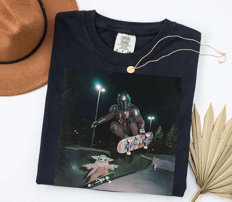 May include: Navy blue long-sleeved t-shirt with a graphic of The Mandalorian and Grogu skateboarding at night. The shirt has a crew neck and a Comfort Colors tag. A gold necklace and a brown hat are also in the image.