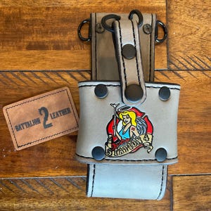 May include: A tan leather accessory with a Station 4 graphic and the text "Battalion 2 Leather." The accessory has black rivets and metal rings. The Station 4 graphic features a woman with angel wings.